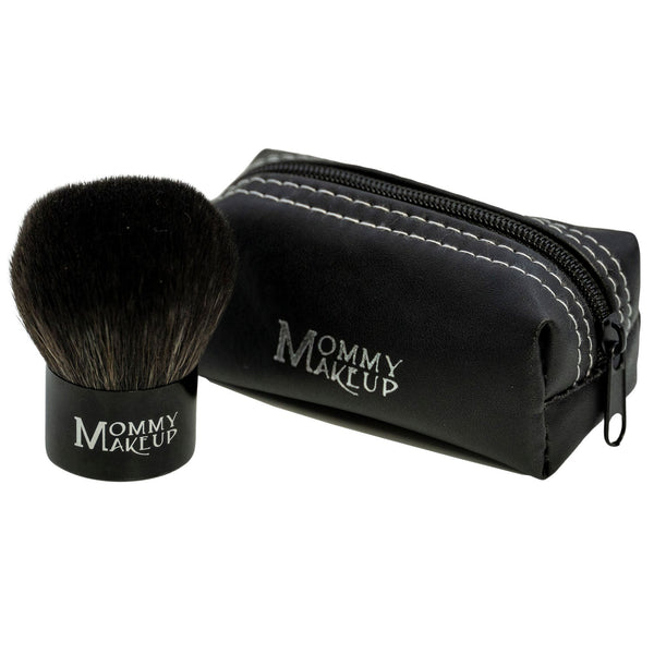 Mommy Makeup Kabuki Brush featuring soft synthetic bristles and a sleek handle, resting on a beige travertine plinth.
