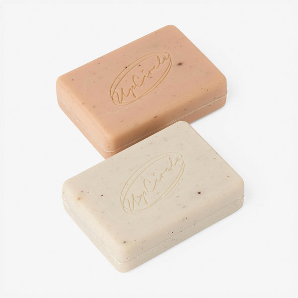 UpCircle Chai Soap Cleansing Duo resting on a textured travertine plinth against raw oatmeal silk, minimal clean aesthetic.