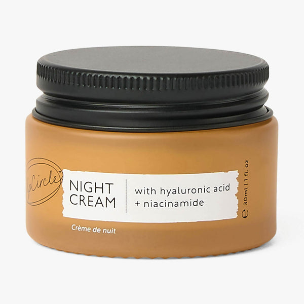 UpCircle Night Cream travel-size glass jar sitting on a textured travertine plinth against a backdrop of raw silk linen.