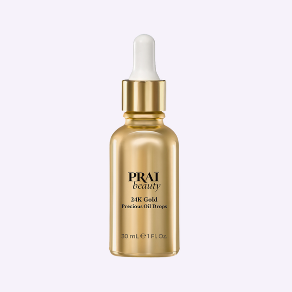 PRAI Beauty 24K Gold Precious Oil Drops glass dropper bottle resting on a textured travertine surface.
